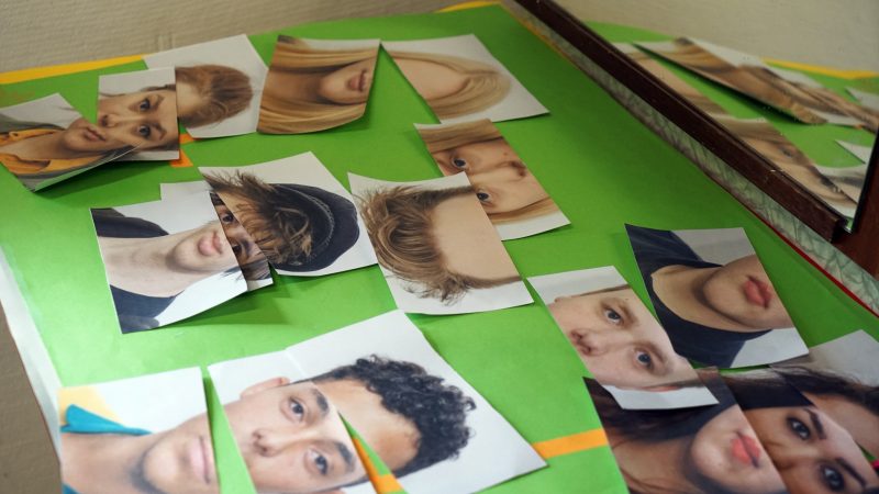 Auf einem Tisch liegen bunte Plakate, das oberste Plakat grün, auf dem horizontal drei Mail zerschnittene Portraits von Jugendlichen liegen. Die einzelnen Ausschnitte der Portraits sind teilweise zusammenhängend, teilweise erbeben sie neu Zusammengesetzte Gesichter.