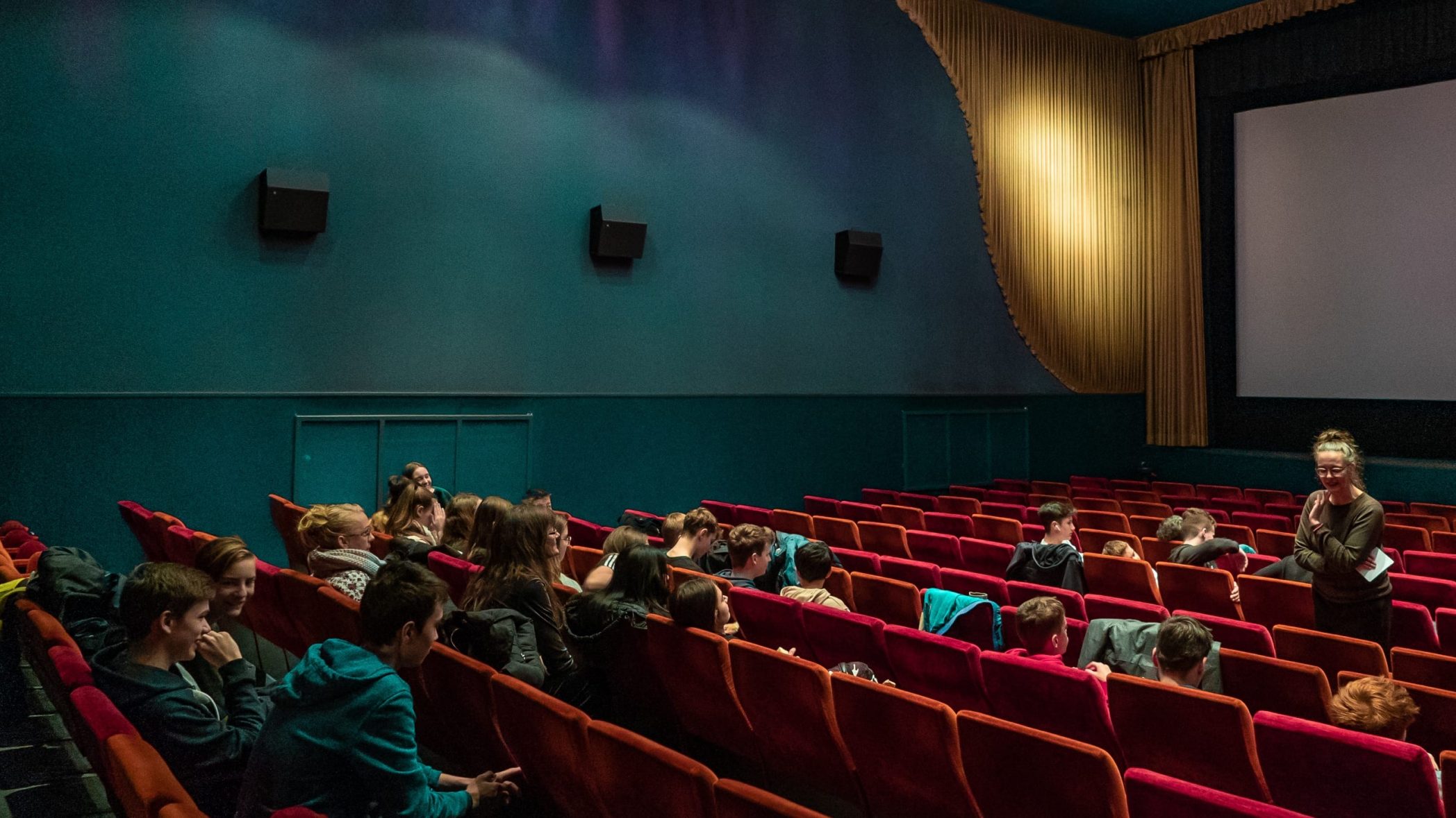 Inneres eines Kinosaals mit roten Kinosesseln und blaugrünen Wänden. Eine weiße Frau mit braunem Langarmshirt, Brille und Dutt steht zwischen den Kinosesseln und etwa 20 Jugendliche, die verteilt in den Sesseln sitzen, teilweise zuhörend, teilweise miteinander beschäftigt, etwas vor.