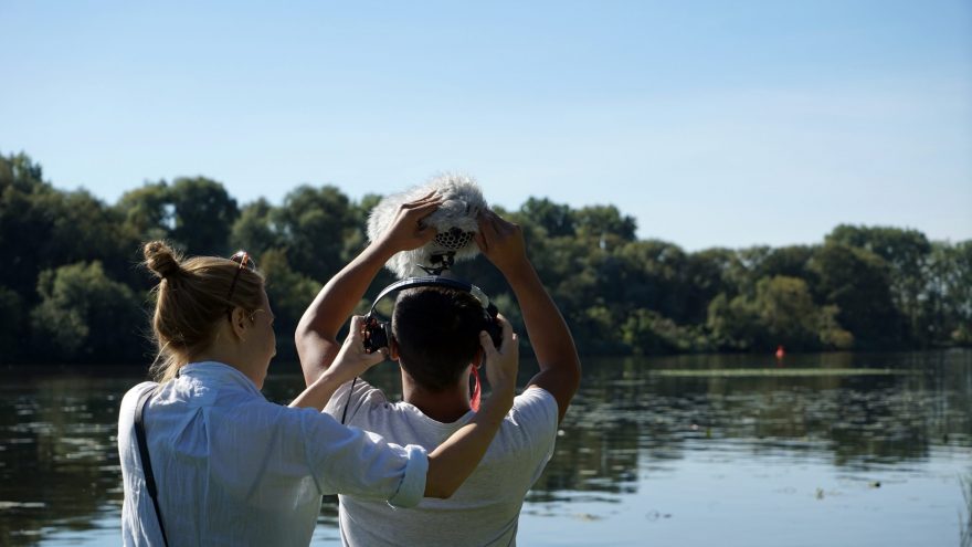 Rückenansicht von zwei jungen Menschen an einem See. Eine Person hält eine Tonangel, die andere Person setzt ihr einen Kopfhörer auf.